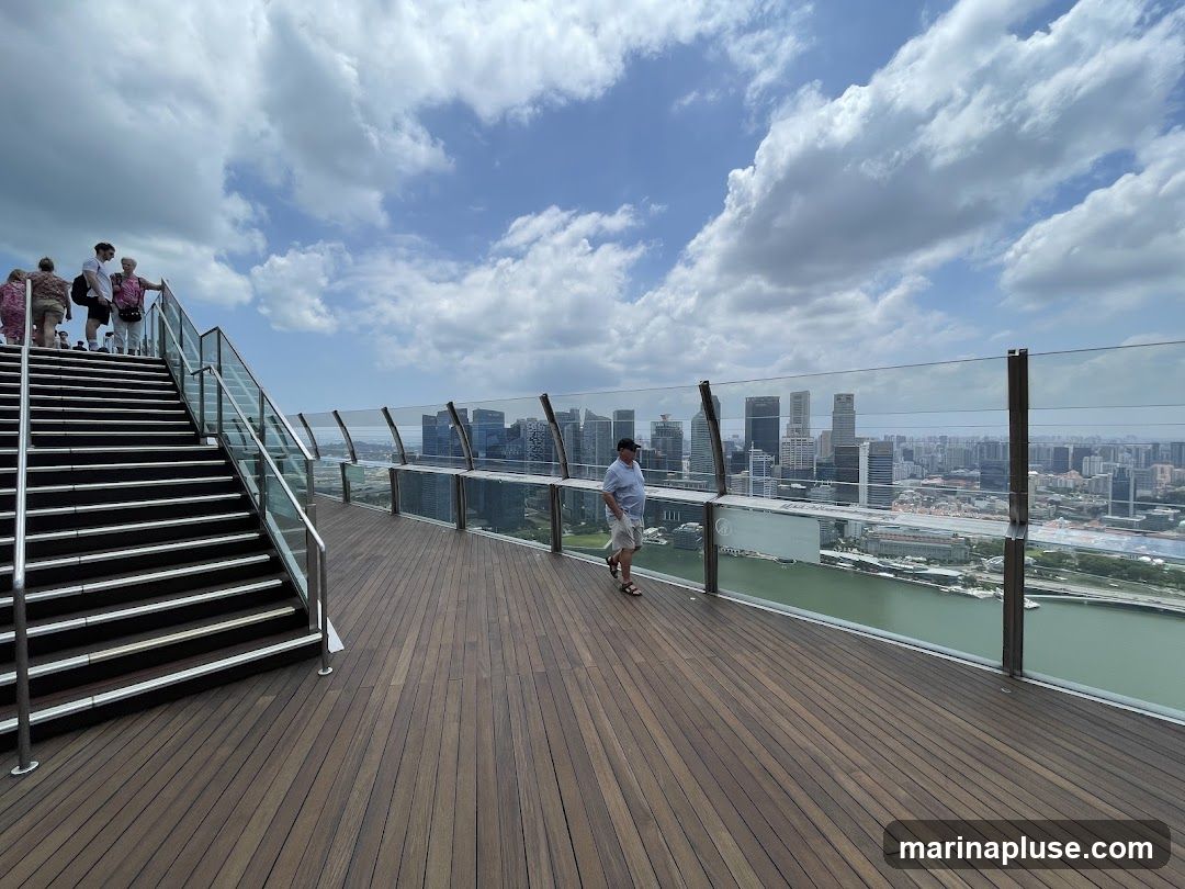 A group of people stand on a wide staircase leading up to an observation deck. The deck has a wooden floor and is bordered by a glass barrier offering a panoramic view of a city skyline and a body of water. A man walks along the deck, looking out at the view. The sky is partly cloudy.