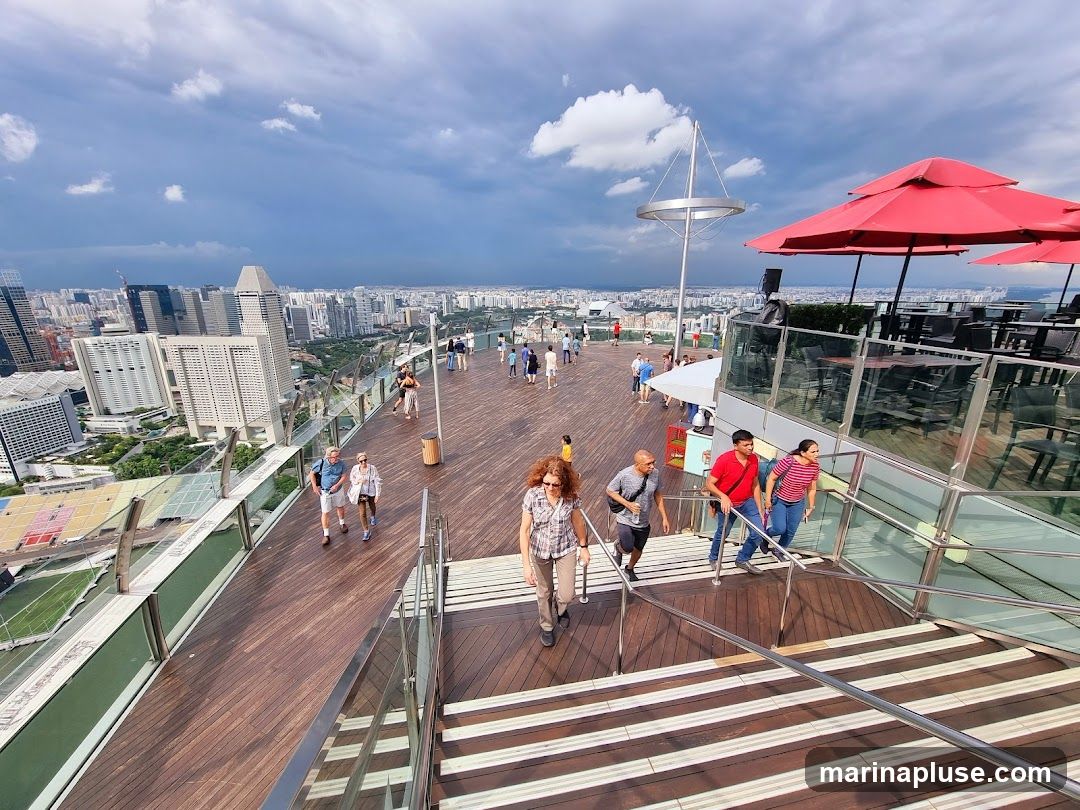 People are walking on a wooden deck on a rooftop observation deck, with a panoramic view of a city skyline under a cloudy sky. There are tables and chairs with red umbrellas visible on the right, suggesting an outdoor dining or lounge area.