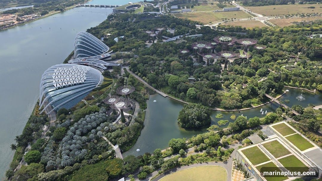 An aerial view of Gardens by the Bay in Singapore, showcasing its iconic Supertree Grove, Cloud Forest, and Flower Dome conservatories. The lush greenery, water features, and modern architectural structures are visible, set against a backdrop of the Singapore Strait and urban landscape.