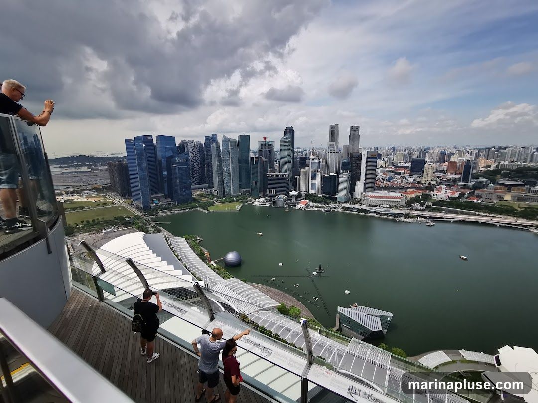 A high-angle shot captures the sprawling Singapore skyline, with numerous skyscrapers dominating the view. A body of water, likely Marina Bay, separates the city from a large, modern architectural structure with a distinctive white, curved roof. Several people are visible on a wooden deck in the foreground, some taking photos and pointing towards the cityscape. The sky is partly cloudy, with dramatic grey clouds contrasting with patches of blue.