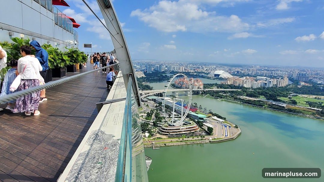 A wide-angle view from a high vantage point, likely an observation deck, overlooking the Singapore skyline. In the foreground, a wooden walkway with people enjoying the view. Lush green plants in planters line the walkway. A prominent cable barrier separates the walkway from the edge. In the mid-ground, the Singapore Flyer Ferris wheel is visible, along with a body of water and surrounding urban landscape. The background shows a dense cityscape under a bright blue sky with scattered clouds.