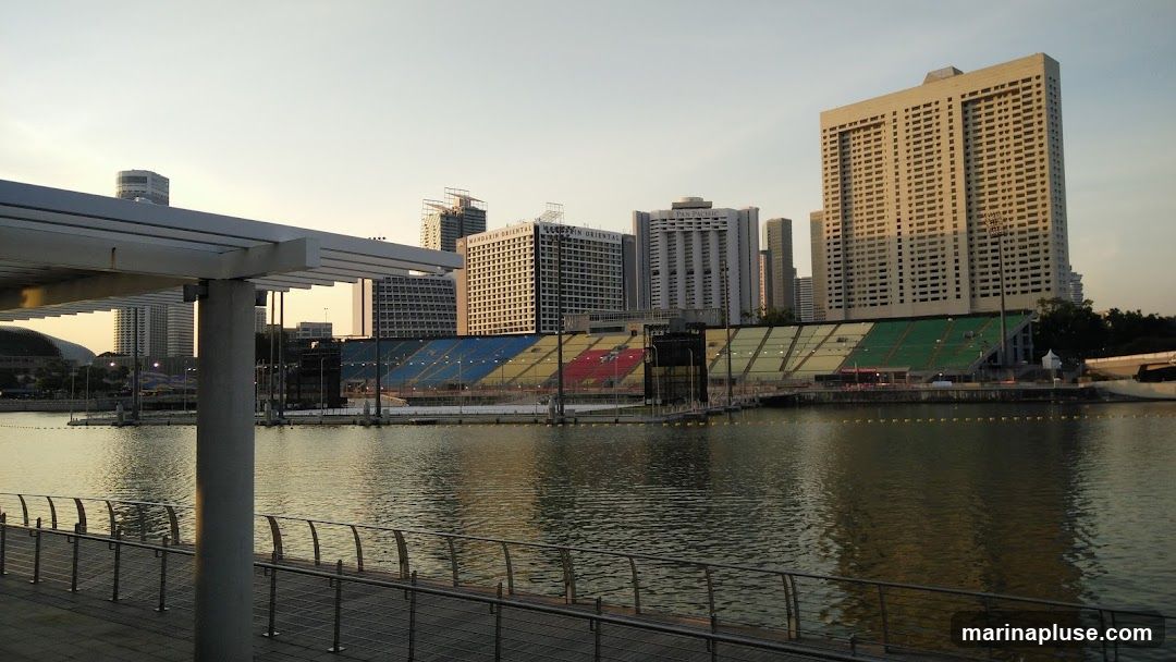 A wide shot of a waterfront area with a cityscape in the background. In the foreground, a walkway with a metal railing overlooks a body of water. Across the water, a stadium with colorful seating (blue, yellow, red, and green) is visible. Behind the stadium, several tall buildings rise into the sky, including buildings labeled 'MANDARIN ORIENTAL' and 'PAN PACIFIC'. The sky is clear and pale, suggesting either early morning or late afternoon light.