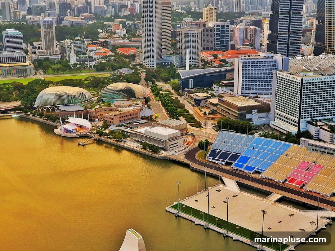 An aerial view captures the bustling cityscape of Singapore, with a focus on the Marina Bay area. Prominent structures include the distinctive durian-shaped Esplanade – Theatres on the Bay, the Marina Mandarin hotel, and a grandstand with colorful seating. The golden hue of the water reflects the urban landscape, creating a vibrant scene. The image showcases a blend of modern architecture and waterfront development.