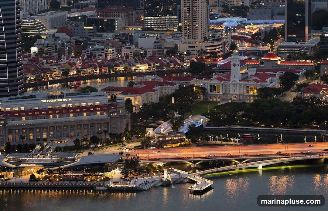 An aerial view of a city skyline at dusk, with a river flowing through it. The Fullerton Hotel is prominently visible, along with other buildings and bridges illuminated by lights. The scene captures the vibrant atmosphere of the city at night.
