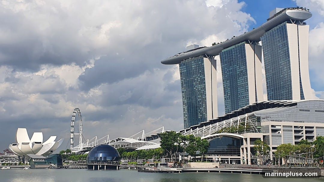 A wide shot of the Marina Bay Sands complex in Singapore under a cloudy sky. The iconic three towers with the SkyPark on top are visible, along with the ArtScience Museum resembling a lotus flower, a geodesic dome structure, and a Ferris wheel in the distance. The waterfront promenade and some greenery are also in view.