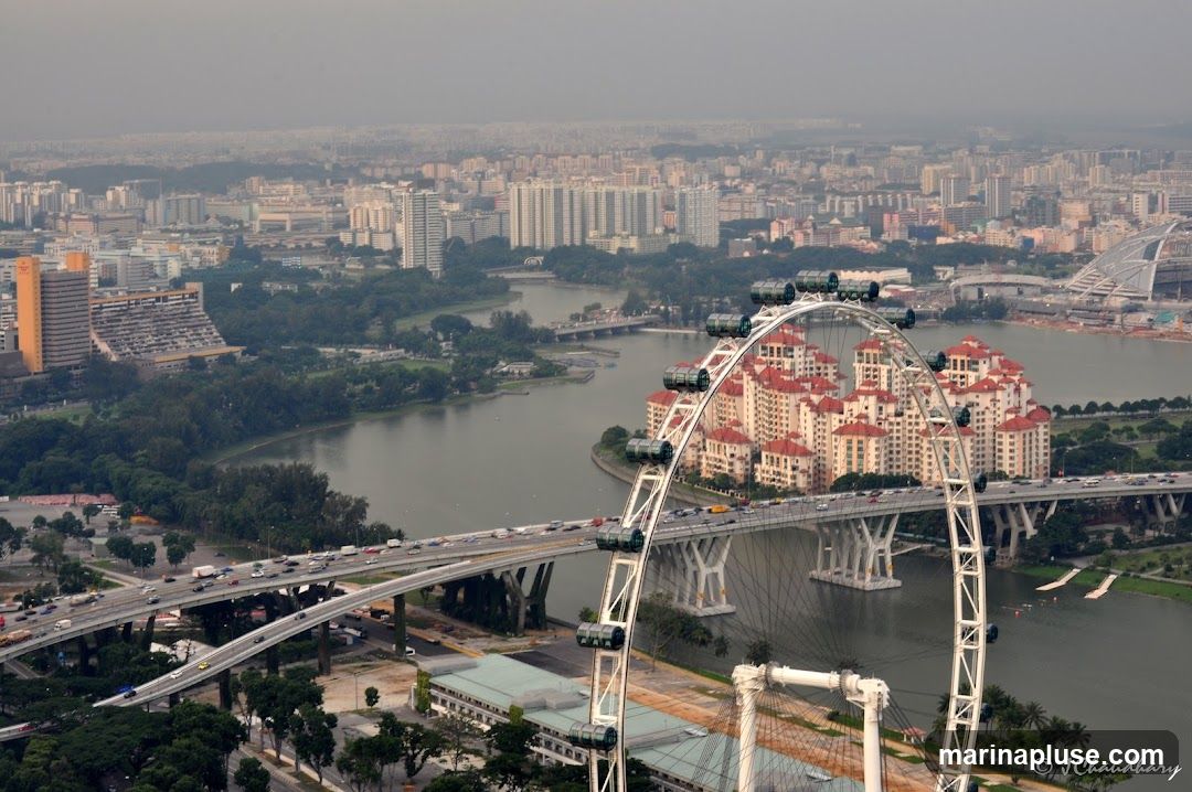 An aerial view of a city skyline, featuring a large Ferris wheel in the foreground. The Ferris wheel's structure is white and prominent, with several gondolas visible. It is situated over a body of water, with a bridge carrying traffic in front of it. In the background, a dense urban landscape with numerous buildings of varying heights stretches towards the horizon under a hazy sky. Residential buildings with red roofs are clustered near the Ferris wheel. The overall scene captures a bustling cityscape with a major landmark.