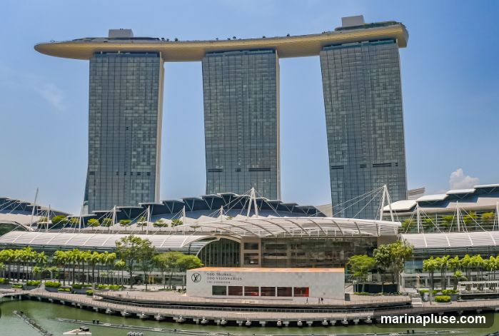 A wide shot of the Marina Bay Sands hotel in Singapore, a resort with three tall towers connected by a sky-high platform. In the foreground, a modern building with the Louis Vuitton logo and text that reads "100 TRUNKS, 100 VISIONARIES, THE EXHIBITION" is visible. The building is situated by a body of water with a promenade in front of it. Lush greenery and palm trees surround the area.