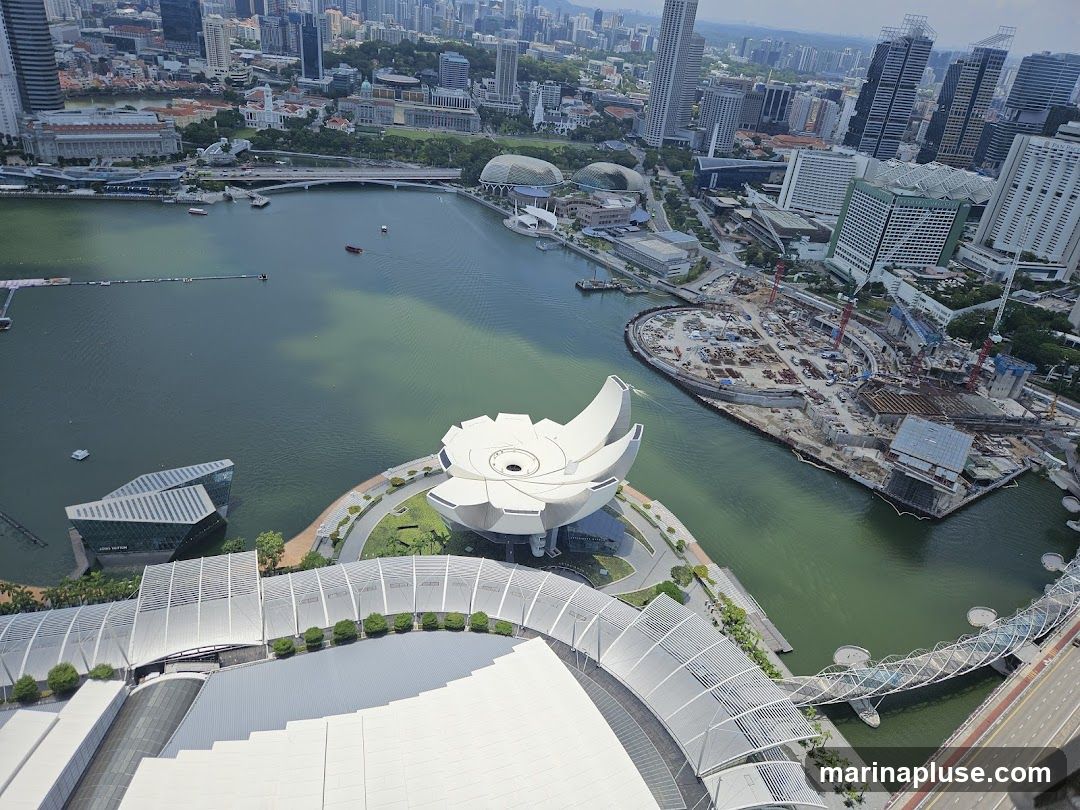 An aerial view of Marina Bay Sands in Singapore, showcasing its iconic lotus-shaped ArtScience Museum and the surrounding cityscape. The image captures the bay with boats, a bridge, and a construction site on an adjacent island. The foreground shows the distinctive architecture of the Marina Bay Sands complex.