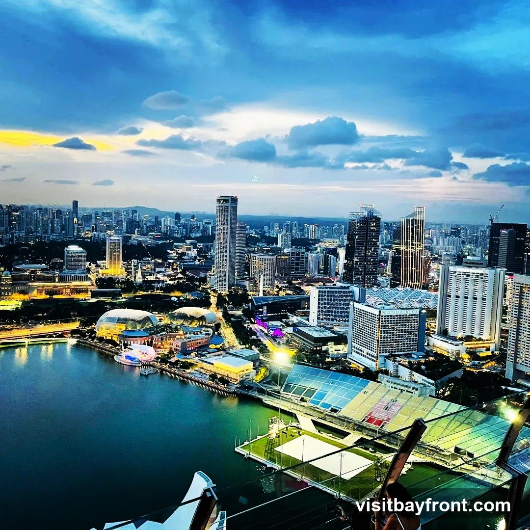 An aerial view of a sprawling cityscape at dusk, with a body of water in the foreground. Numerous skyscrapers and buildings are illuminated, creating a vibrant urban landscape. The sky is filled with dramatic clouds, with a hint of yellow and orange from the setting sun on the horizon. In the middle ground, distinctive domed structures and a modern stadium-like building with colorful panels are visible. The overall scene captures the energy and beauty of a city as night falls.