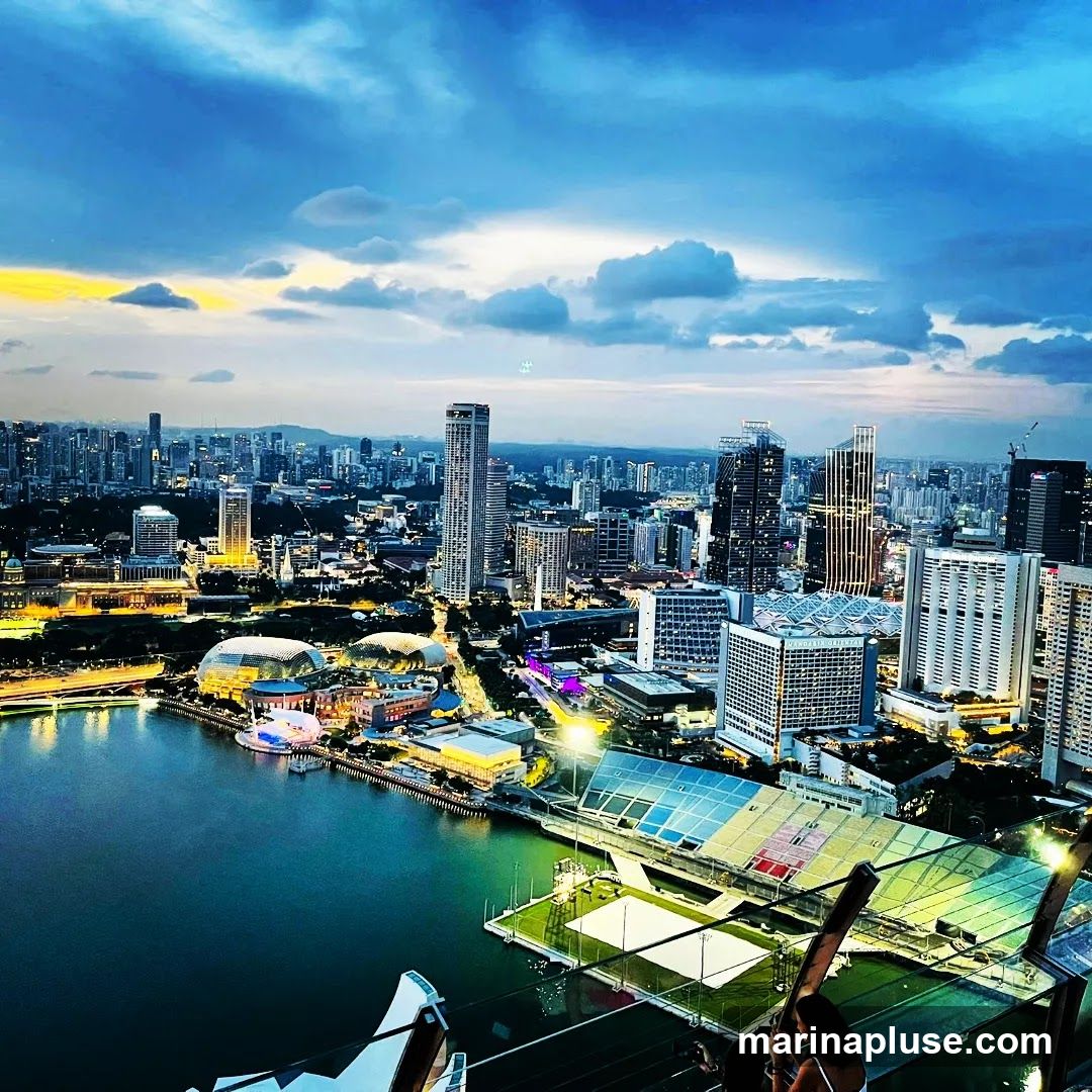 An aerial view of a sprawling cityscape at dusk, with a body of water in the foreground. Numerous skyscrapers and buildings are illuminated, creating a vibrant urban landscape. The sky is filled with dramatic clouds, with a hint of yellow and orange from the setting sun on the horizon. In the middle ground, distinctive domed structures and a modern stadium-like building with colorful panels are visible. The overall scene captures the energy and beauty of a city as night falls.
