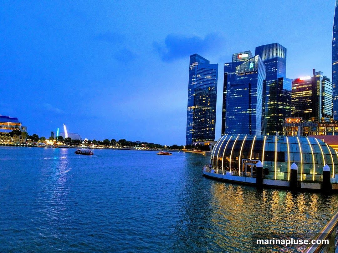 A wide shot of a city skyline at dusk, with tall modern buildings illuminated by lights. In the foreground, a body of water reflects the lights of the city and a unique, dome-shaped building with glowing vertical lines. Several boats are visible on the water. The sky is a deep blue, with some clouds.