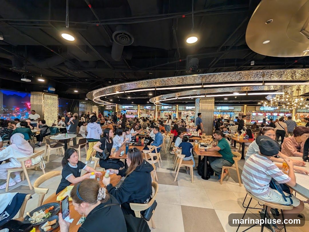 A bustling food court filled with people dining at various tables. The ceiling is dark with exposed pipes and lighting fixtures. A decorative, illuminated circular structure hangs above the seating area. In the background, multiple food stalls are visible with signage, and customers are queuing and ordering food. The overall atmosphere is lively and crowded.