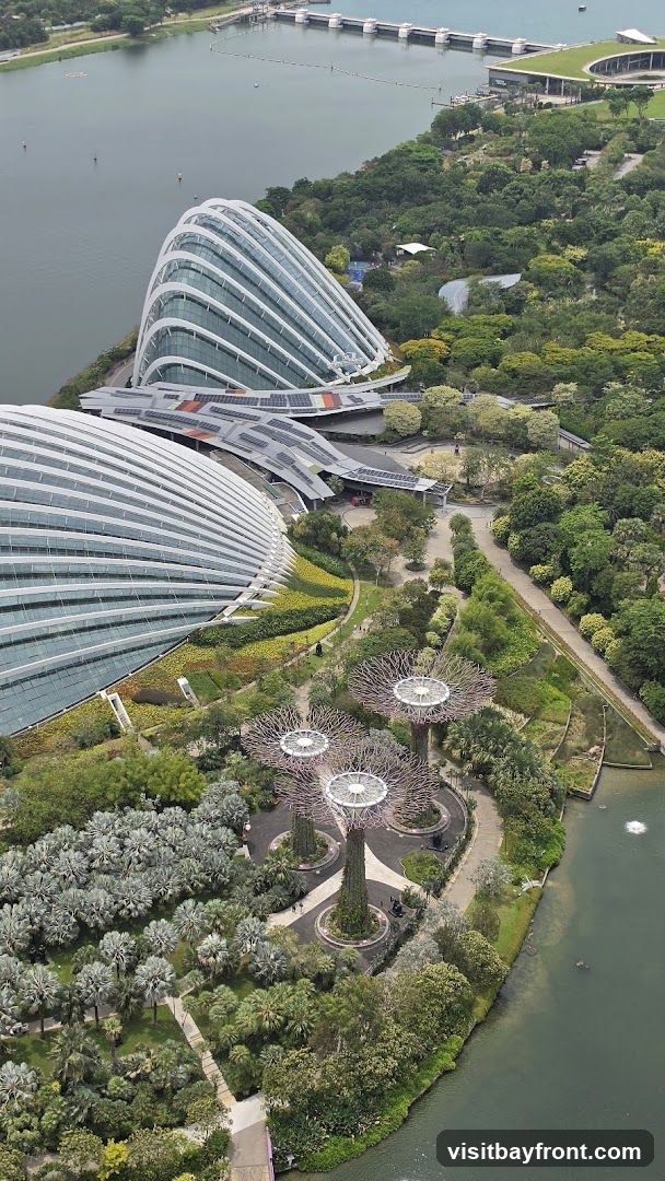 An aerial view of Gardens by the Bay in Singapore, showcasing the iconic Supertree Grove and the large conservatories. The image captures the lush greenery, pathways, and the waterfront, with a dam structure visible in the background.