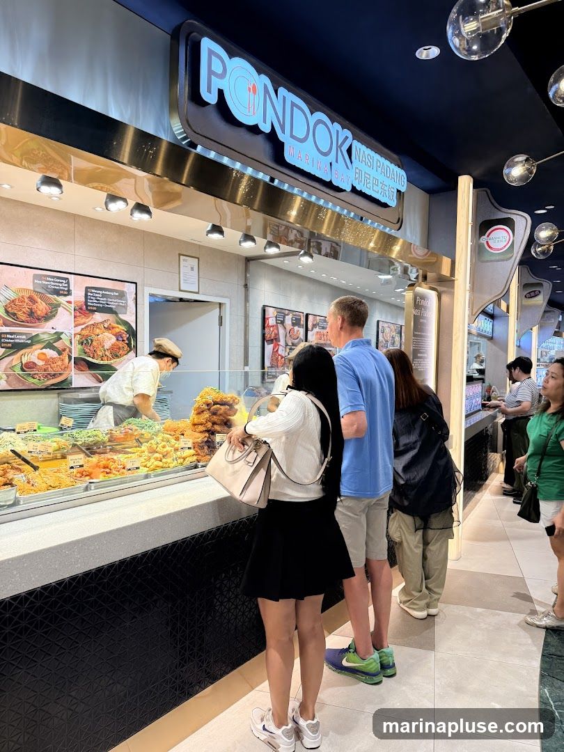 A food stall counter with various dishes displayed behind a glass partition. People are queuing to order. The stall has a prominent sign that reads "PONDOK MARINA BAY NASI PADANG". There are also menu boards with pictures of food items and prices.