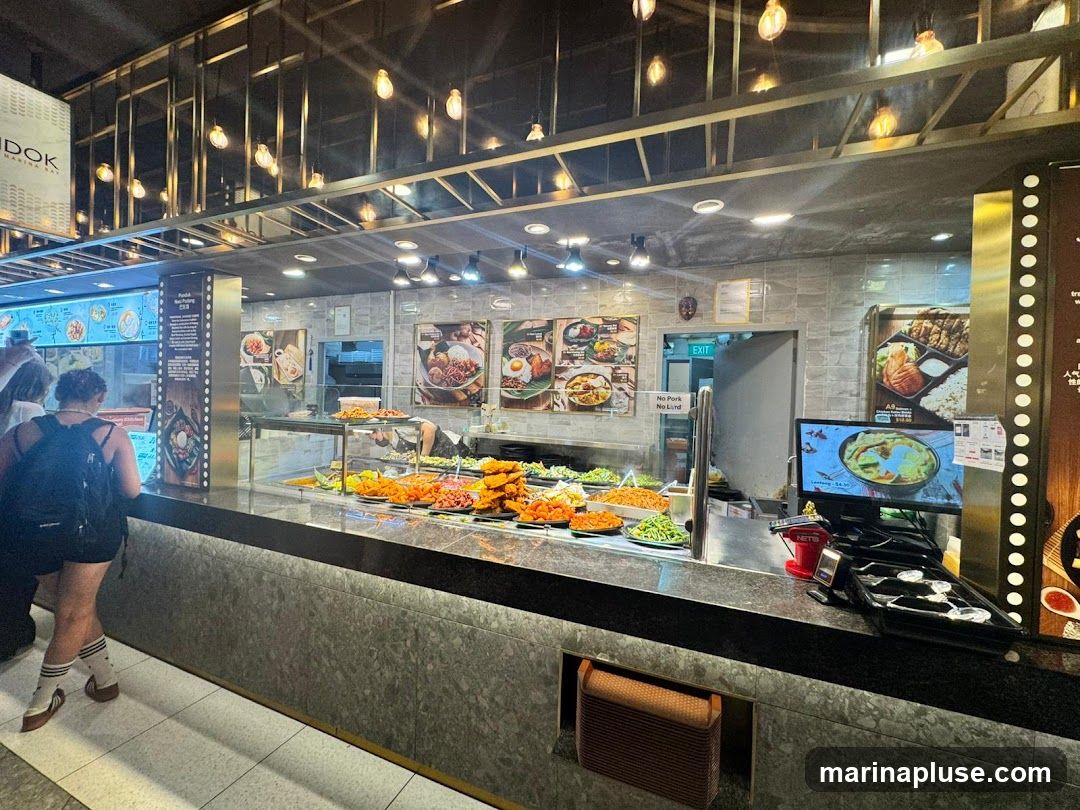 A food stall counter with various dishes displayed in trays. There are pictures of food on the wall and a digital menu screen. A person is standing at the counter, looking at the menu.