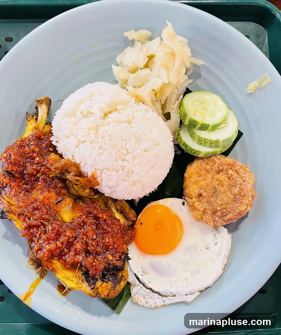 A plate of Nasi Lemak with a fried egg, fried chicken, rice, pickled vegetables, cucumber slices, and a fried patty.