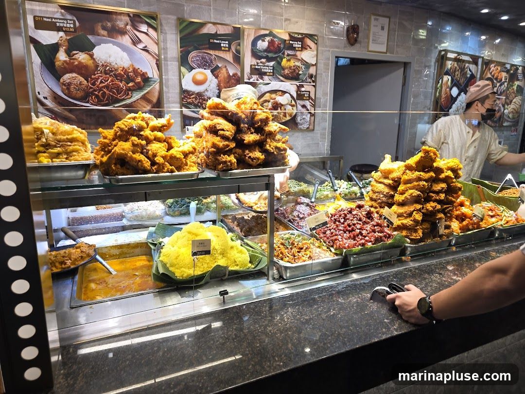 A food stall display featuring a variety of dishes, including fried chicken, curries, rice, and vegetables. A staff member in the background is serving customers. Several framed pictures of menu items are visible on the wall.