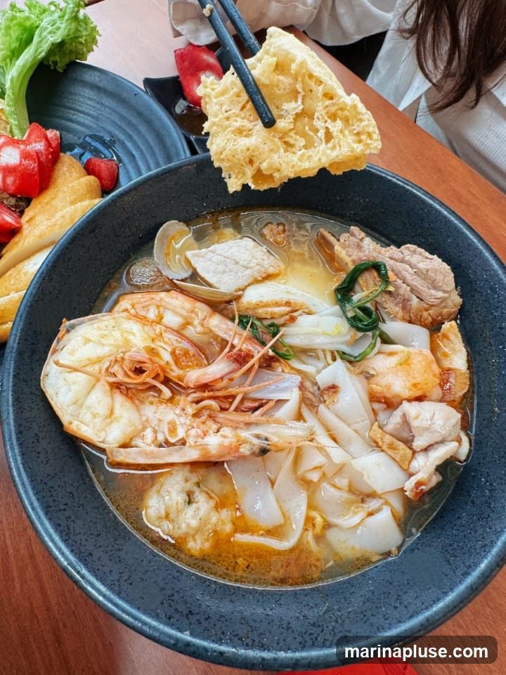 A close-up shot of a bowl of noodle soup with large prawns, clams, sliced meat, and flat noodles in a savory broth. A piece of fried dough is being lifted out of the bowl with chopsticks. In the background, there are side dishes of fruit and lettuce.