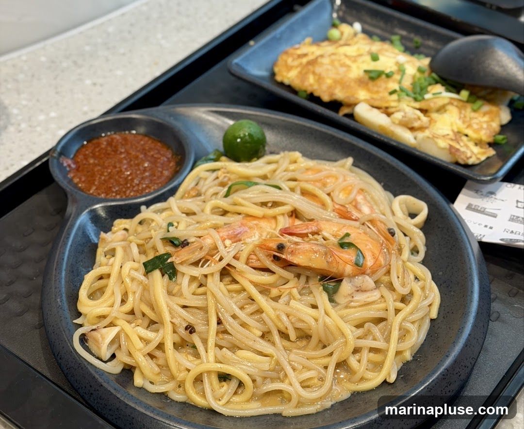 A close-up shot of a plate of noodles with shrimp and calamari, served with a side of chili sauce and a lime wedge. In the background, another plate holds an omelette garnished with chopped green onions.