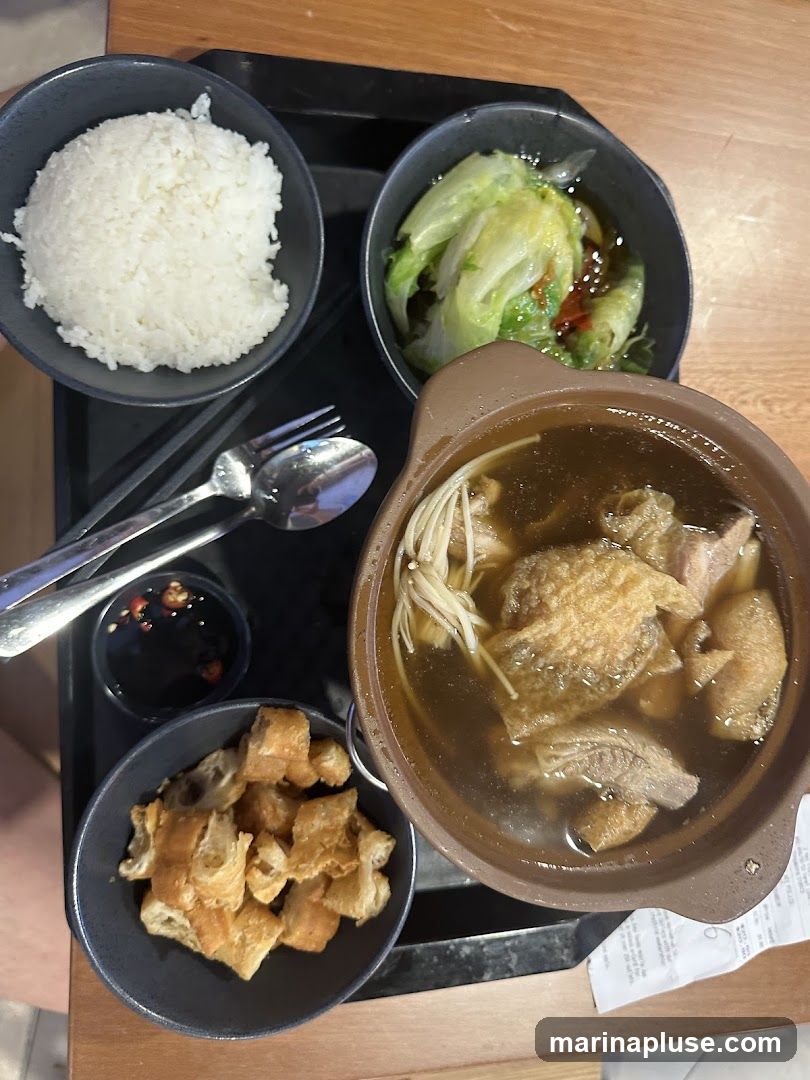 A top-down view of a meal served on a black tray. The meal includes a bowl of white rice, a bowl of braised lettuce, a bowl of fried dough sticks (youtiao), and a clay pot of Bak Kut Teh with meat and enoki mushrooms. A small dish of chili and soy sauce is also visible, along with chopsticks and a spoon and fork. A receipt is partially visible at the bottom right.