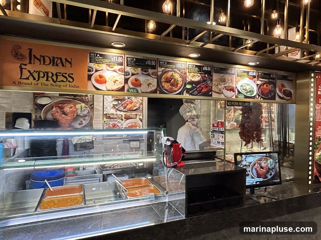 A food counter with a menu board displaying various Indian food sets and dishes. The menu includes options like Set A, Set B, Set C, Set D, Set E, and Set F, with prices ranging from $10.90 to $16.90. Some dishes are labeled as VEG, and there is a prominent "NO PORK NO LARD" sign. Food items are displayed in trays behind a glass counter, and skewers of meat are hanging. A digital display shows a picture of a chicken dish.