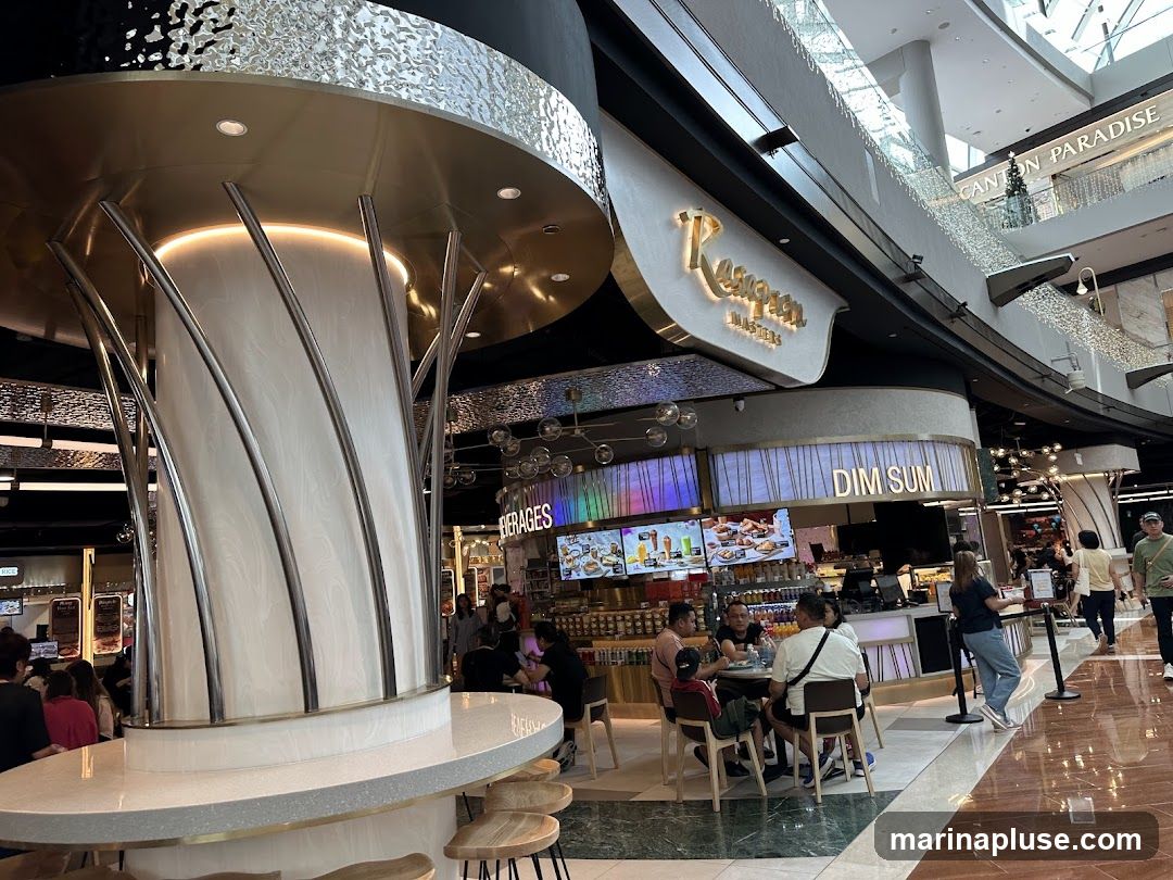 A bustling food court scene with various food stalls. In the foreground, a modern, white pillar with metallic accents stands prominently. To the right, a stall with a sign reading "DIM SUM" is visible, displaying colorful images of food on a screen. Further back, another stall has a sign for "BEVERAGES" and a menu board with pictures of drinks and food. People are seated at tables, eating and socializing. The overall ambiance is lively and commercial, typical of a shopping mall food court.