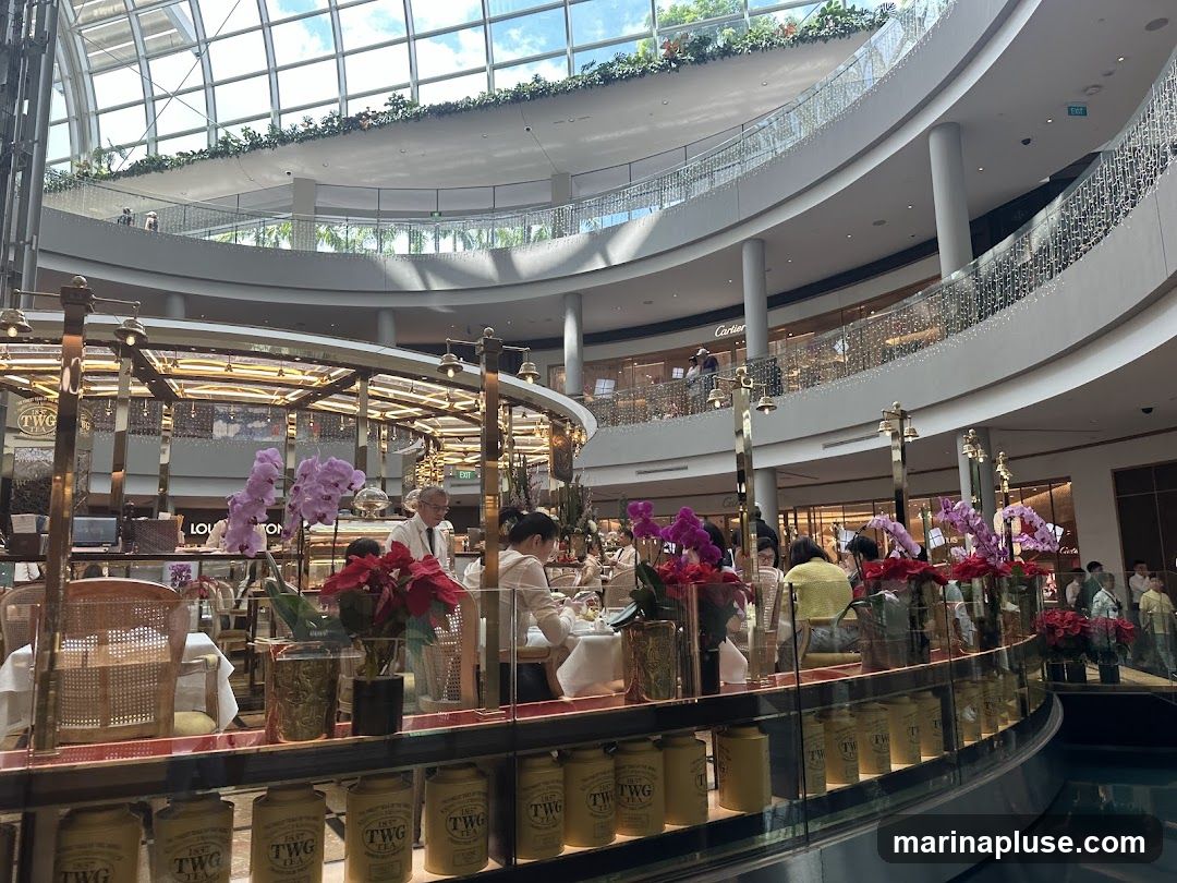An interior view of a cafe or restaurant located within a large, multi-level shopping mall. The cafe features a prominent display of floral arrangements, including orchids and poinsettias. Patrons are seated at tables, some of which are set with white tablecloths. The cafe's branding, "TWG Tea," is visible on numerous canisters lining a display shelf. In the background, other retail stores are visible on different levels of the mall, including Cartier and Louis Vuitton. The mall's architecture includes a large glass ceiling and curved balconies adorned with decorative lights.
