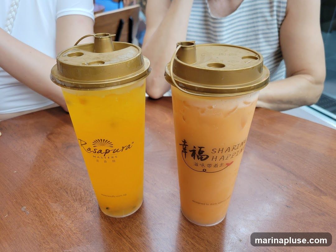 Two plastic cups with golden lids filled with beverages sit on a wooden table. The cup on the left contains a yellow, pulpy drink, and the cup on the right contains an orange, creamy drink. Both cups have logos and text printed on them. The logos indicate the brand names "Rasapura MASTERS" and "SHARING HAPPINESS".