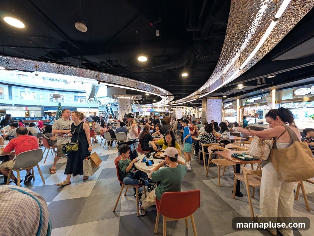 A bustling food court filled with people dining at tables and walking around. The ceiling features modern, textured lighting fixtures. Various food stalls are visible in the background, with some displaying menus and food items.