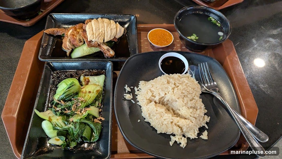 A tray of Hainanese chicken rice with roasted chicken, bok choy, rice, soup, and chili sauce.