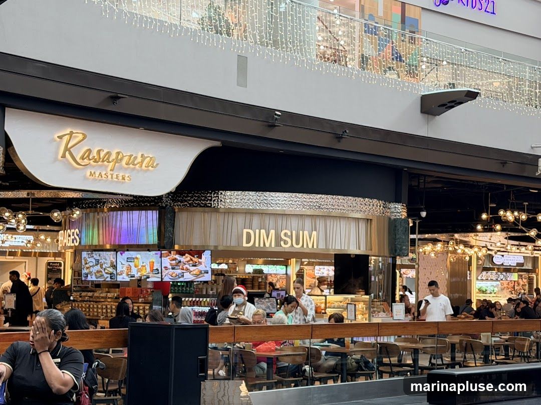 A busy food court area with a restaurant called Rasapura Masters specializing in Dim Sum. People are seated at tables, and food displays are visible.