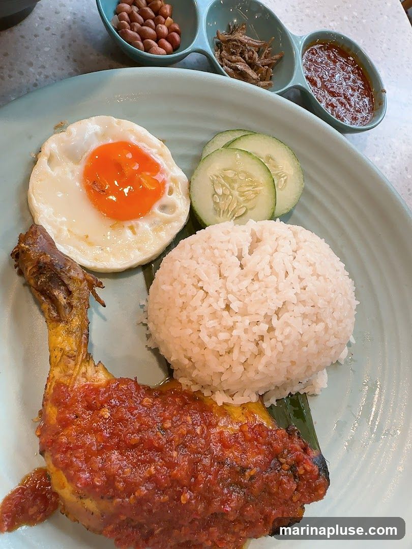 A plate of Nasi Lemak featuring a fried egg, a mound of white rice, a piece of chicken coated in chili sauce, sliced cucumbers, and three small dishes containing peanuts, anchovies, and sambal.