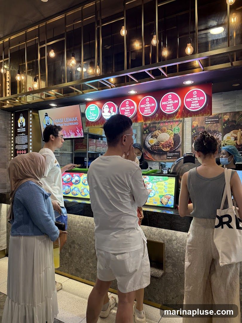 People are standing in line at a food stall counter. Above the counter, there is a screen displaying "NASI LEMAK VAM TALIWANG" with graphics of flames. To the right, there are circular signs indicating "THE MICHELIN GUIDE" for the years 2021, 2022, 2023, and 2024, along with a "HALAL" certification. Menus with food images and prices are visible on the counter.