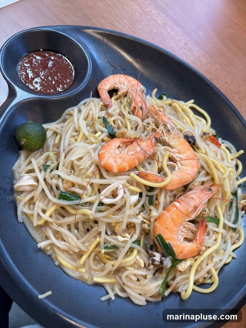 A close-up shot of a plate of stir-fried noodles with shrimp, squid, and green onions. A small dish of chili sauce and a lime wedge are on the side.