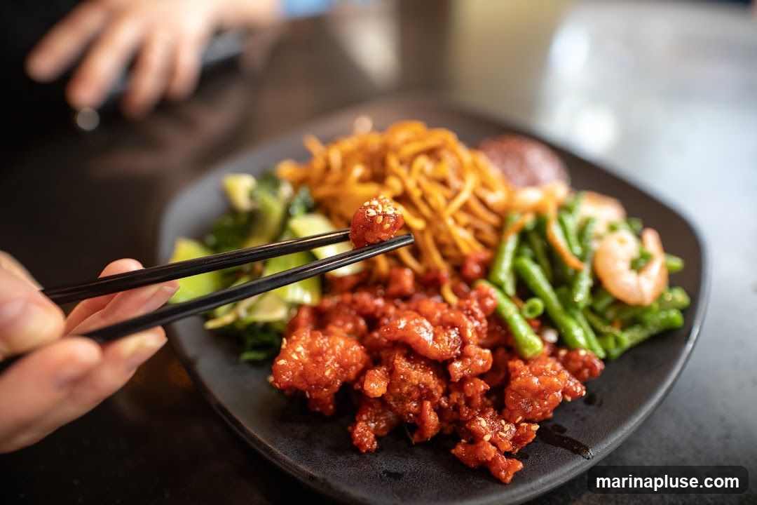 A close-up shot of a dark plate filled with various Asian dishes. In the foreground, a pair of black chopsticks is lifting a piece of sesame-seed-covered meat glazed in a red sauce. The plate also contains stir-fried noodles, green beans with shrimp, and leafy green vegetables. The background is blurred, showing a person's hands and a dark tabletop.