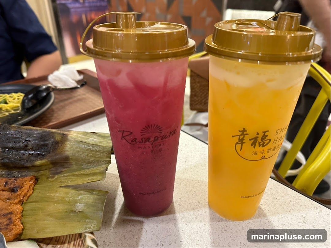 Two tall plastic cups with golden lids, one filled with a pinkish-red drink and the other with a yellow drink, sit on a speckled countertop. The pink drink has a logo that reads "Rasapuri MASTERS" and a website address. The yellow drink has a logo with Chinese characters and the text "SHANG HAI" and "designed to drink". In the foreground, a piece of food wrapped in banana leaves and grilled is visible. In the background, a plate of noodles and a person in a blue shirt can be seen.