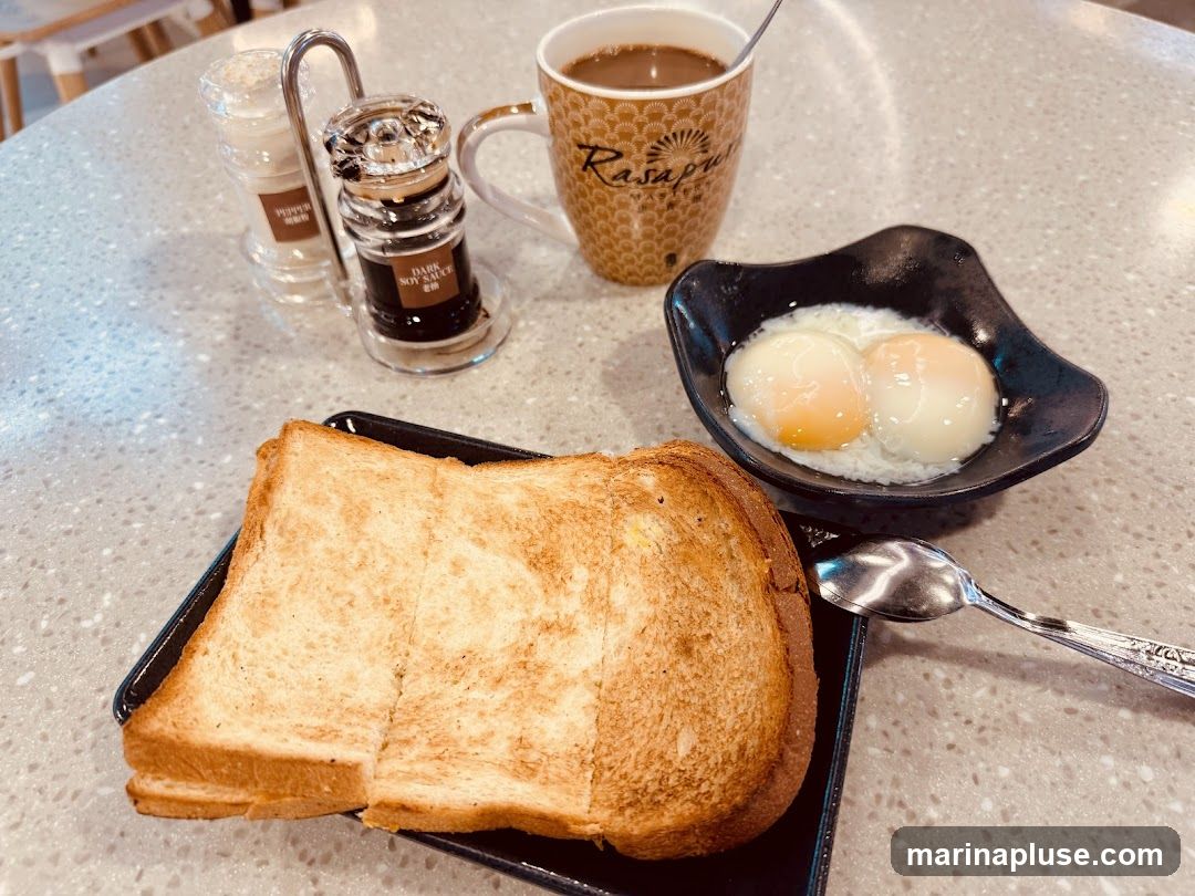 A breakfast set featuring toast, soft-boiled eggs, and coffee. The toast is golden brown and appears to be buttered. Two soft-boiled eggs are served in a black bowl with soy sauce and pepper shakers in the background. A mug with the logo "Rasapuri" is filled with coffee.