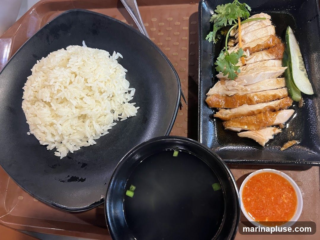 A plate of Hainanese chicken rice is served with sliced chicken, a mound of white rice, a small bowl of soup, and a side of chili sauce. The chicken is glazed and appears tender, garnished with cilantro. Slices of cucumber are also present on the plate. The rice is fluffy and white. The soup is dark and garnished with green onions. The chili sauce is a vibrant orange-red.