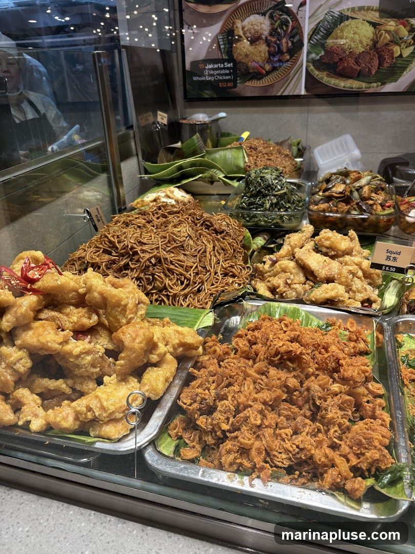 A display of various prepared foods, including fried items, noodles, and vegetable dishes, arranged in metal trays lined with banana leaves. A sign indicates "Squid" for $6.50. In the background, a menu board shows a "Jakarta Set" for $12.80. A person in a white apron is visible behind a glass partition.