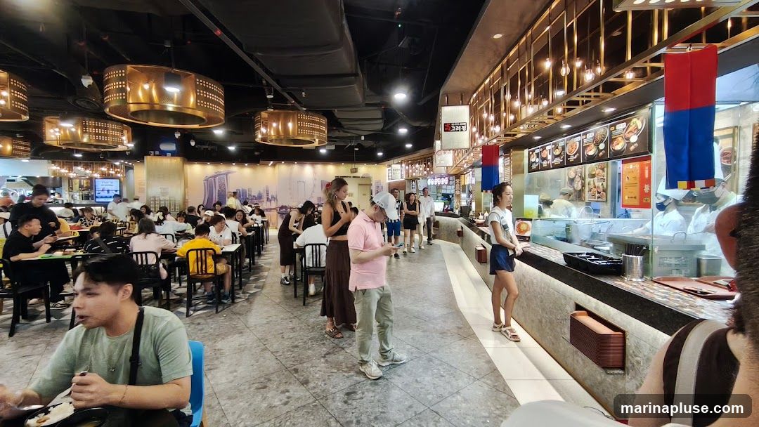 A bustling food court with many patrons seated at tables and queuing at food stalls. The stalls feature illuminated menus with images of dishes. The decor includes modern pendant lights and a mural depicting a city skyline.