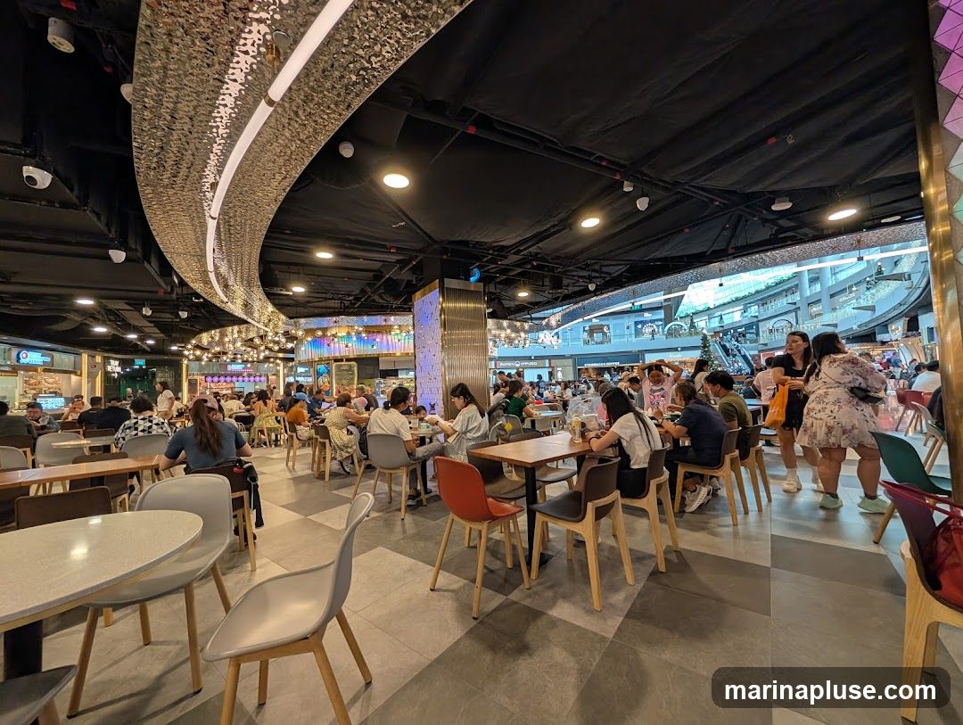 A bustling indoor food court with many patrons seated at tables and dining. The ceiling features a decorative, curved metallic structure with lights, and the floor is tiled in a checkered pattern. In the background, escalators and shops are visible within a larger mall setting.