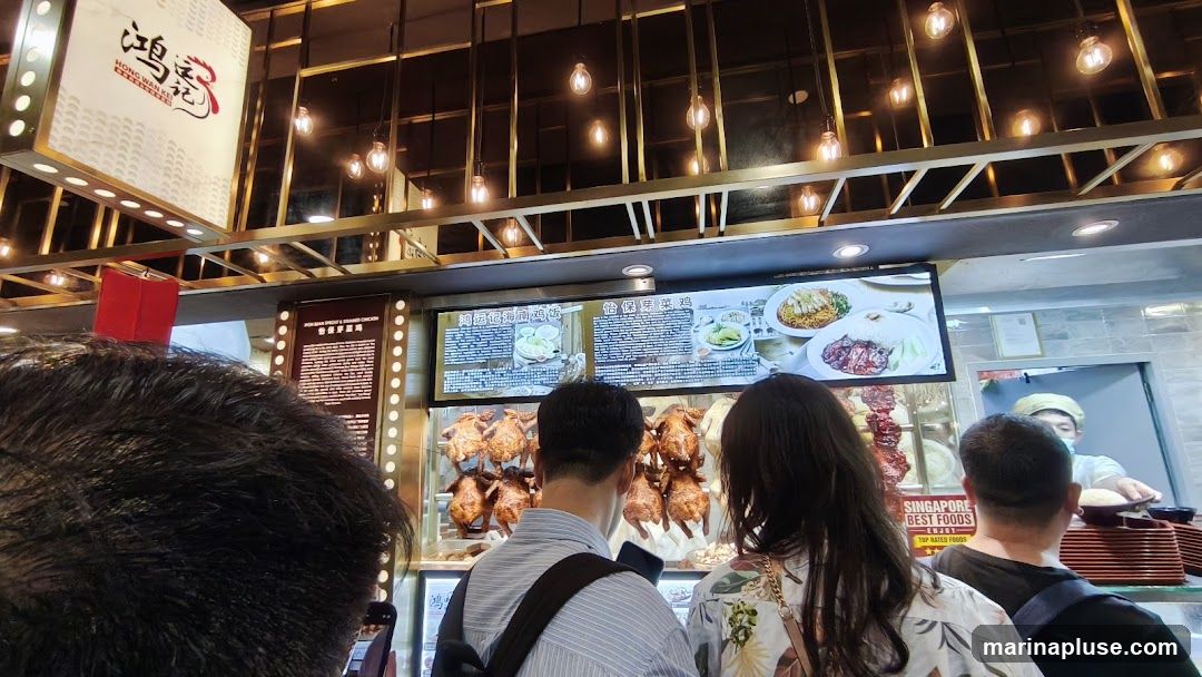 A view of a restaurant counter with several roasted chickens hanging. A digital menu board displays food items and prices. Customers are standing in line, looking at the menu and the food. A staff member is visible behind the counter, serving food. The restaurant has a modern interior with hanging lights.
