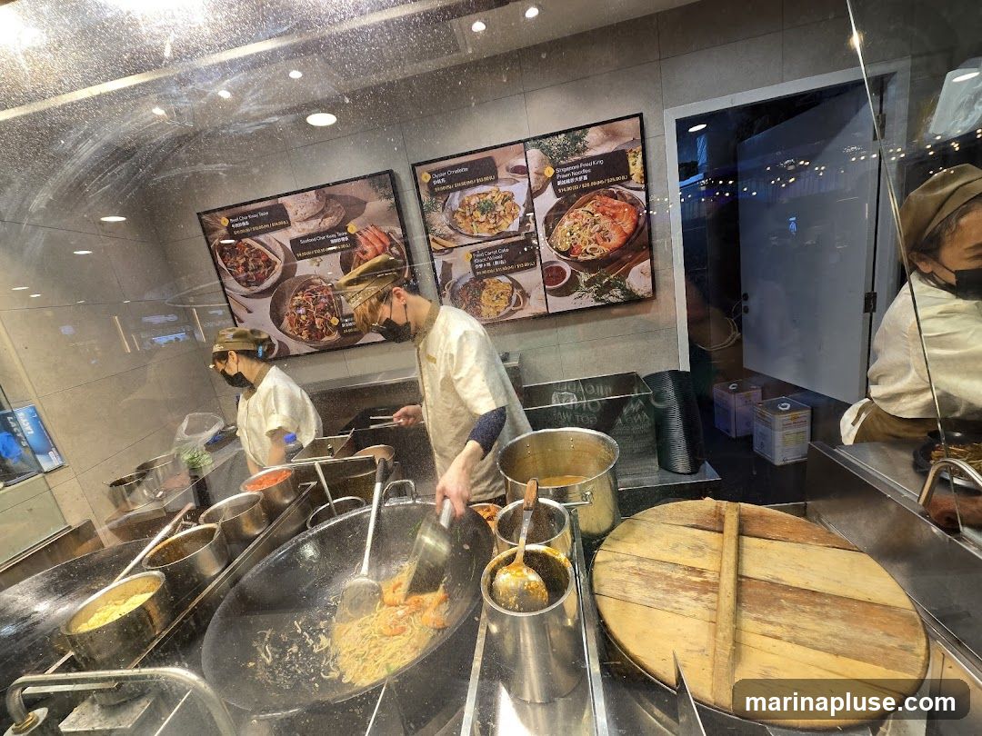 Two chefs in uniform and masks are working in a kitchen. One chef is stir-frying noodles with shrimp in a large wok, while the other is preparing ingredients. Several pots and containers are visible on the counter. Three framed pictures of food dishes are displayed on the wall above the chefs. A large wooden lid covers a pot in the foreground.