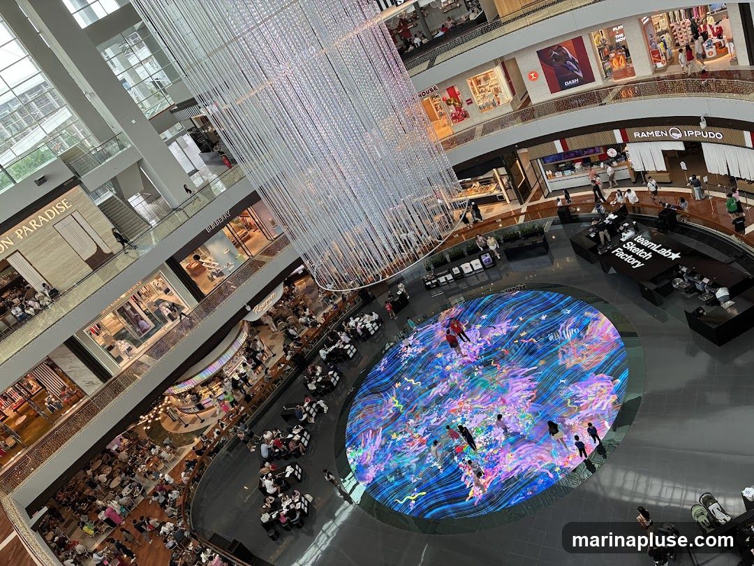 An aerial view of the interior of a multi-level shopping mall. A large, circular digital art display is the centerpiece of the atrium, showing vibrant, flowing colors and shapes. People are seen interacting with the display and walking around the mall. Several shops with visible signage, including 'PARADISE', 'BURBERRY', and 'RAMEN IPPUDO', are located on different levels. A large, ornate chandelier hangs from the ceiling. The mall appears to be busy with shoppers.