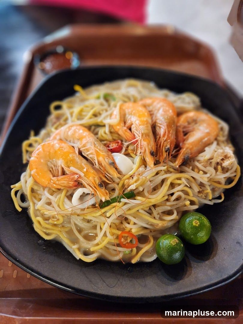 A close-up shot of a plate of noodles topped with several large shrimp, bean sprouts, and chili slices. Two small green limes are placed on the side of the plate. The dish is served on a black plate, which is on a brown tray. The background is blurred, showing hints of a pink garment and a dark object.