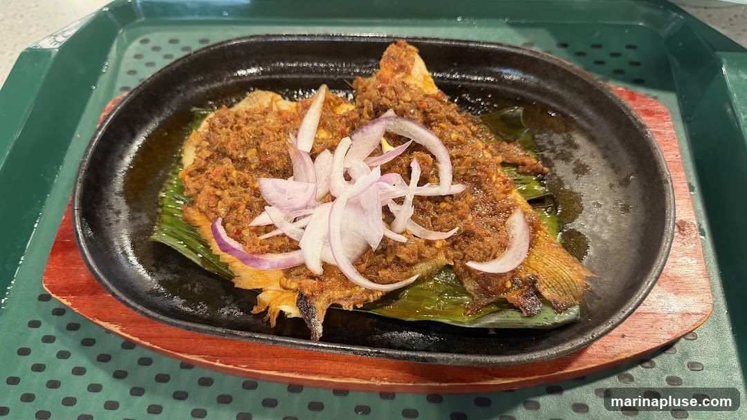 A whole fish, likely fried or grilled, is served on a banana leaf in a black cast-iron skillet. The fish is generously topped with a spicy, reddish-brown sauce and garnished with thinly sliced red onions. The skillet rests on a wooden base, which is placed on a green tray with a textured surface.