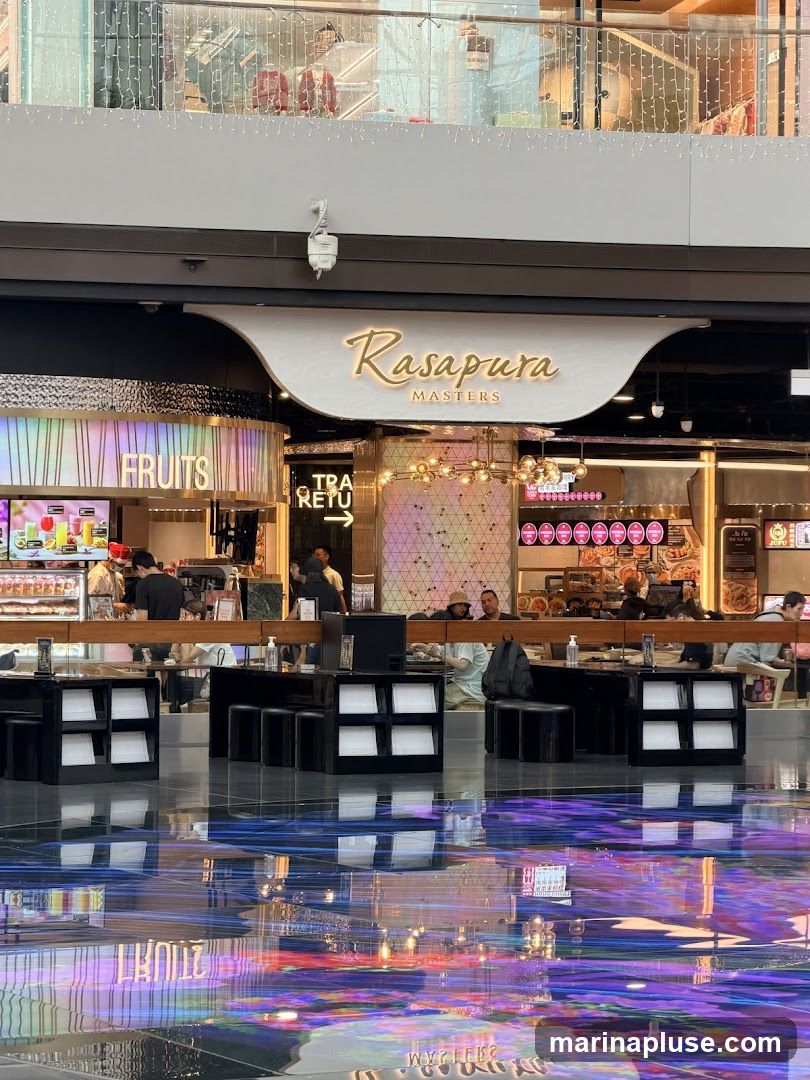 A view of a food court area with tables and chairs in the foreground, and various food stalls in the background. The signage for "RASAPURA MASTERS" is prominently displayed above one of the stalls. Another stall has "FRUITS" written on its facade. The floor has a reflective surface with colorful light patterns.