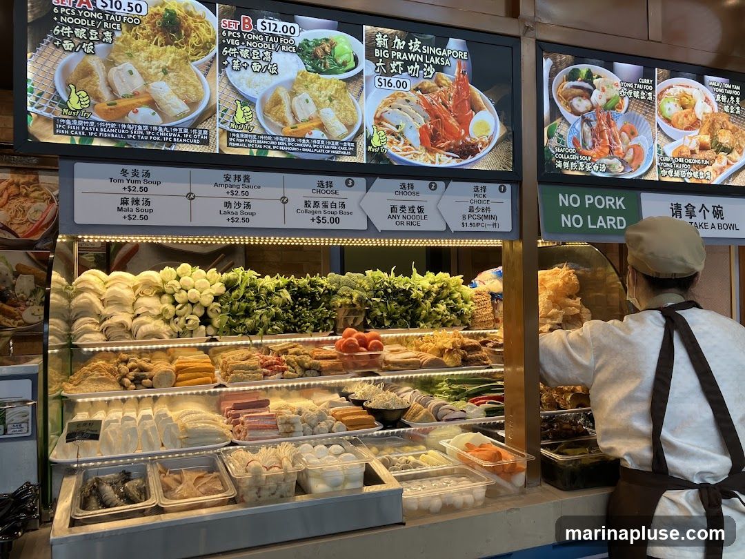 A food stall with a wide variety of fresh ingredients displayed in refrigerated cases. Ingredients include various types of tofu, vegetables, seafood, and processed meats. Above the display are digital menus showing different meal sets and soup bases. A staff member wearing an apron and cap is visible behind the counter. A sign reads "NO PORK NO LARD".