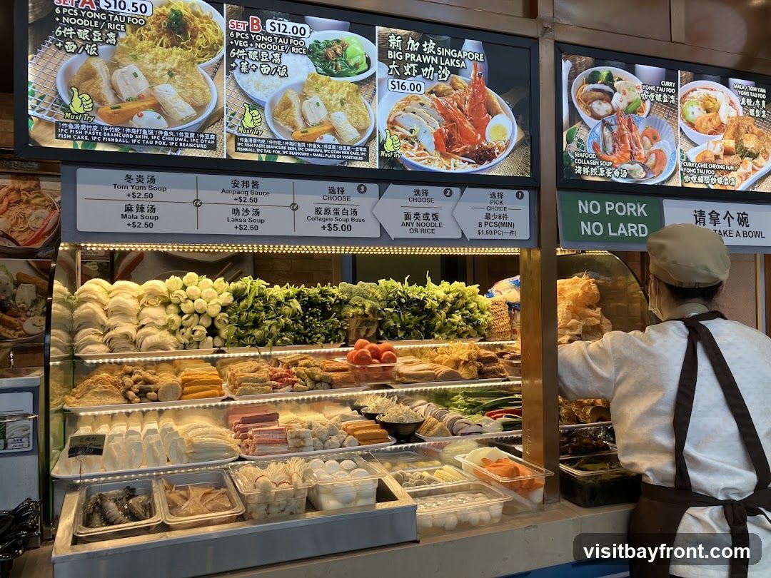 A food stall with a wide variety of fresh ingredients displayed in refrigerated cases. Ingredients include various types of tofu, vegetables, seafood, and processed meats. Above the display are digital menus showing different meal sets and soup bases. A staff member wearing an apron and cap is visible behind the counter. A sign reads "NO PORK NO LARD".
