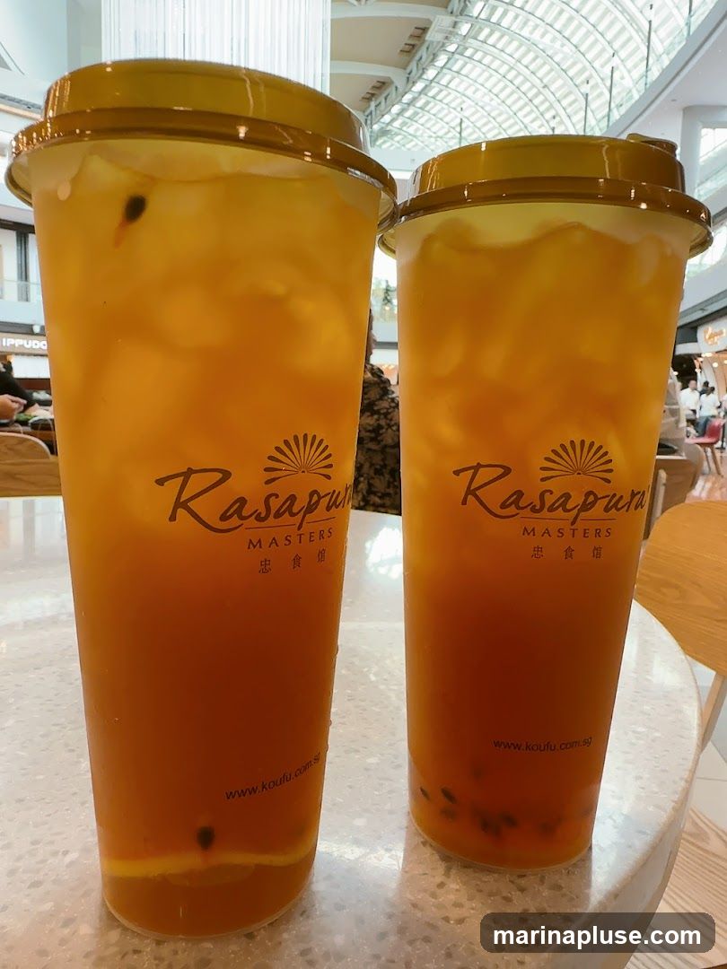 Two large plastic cups filled with a vibrant orange passion fruit drink, complete with ice, passion fruit seeds, and slices of citrus fruit at the bottom. The cups have yellow lids and feature the logo "Rasapura MASTERS" with Chinese characters below, and the website "www.koufu.com.sg" printed near the base. The drinks are placed on a speckled white table, with a blurred background showing a shopping mall interior with a modern, arched ceiling.